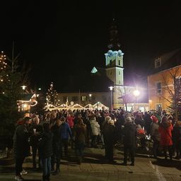 In der Nacht versammeln sich eine Menschenmenge auf einem Stadtplatz, geschmückt mit Weihnachtsbäumen und festlichen Lichtern, vor einem hohen Turm.