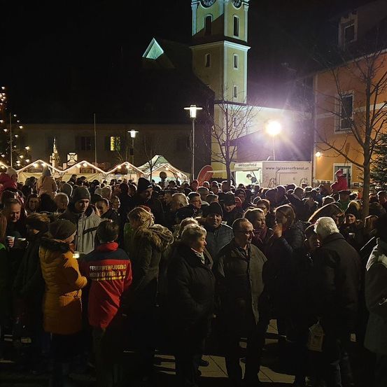 Eine Menschenmenge versammelt sich bei Nacht, in der Nähe eines Glockenturms, mit festlichen Lichtern, Zelten und Bäumen. Viele Menschen tragen Winterkleidung und einige halten Tassen.