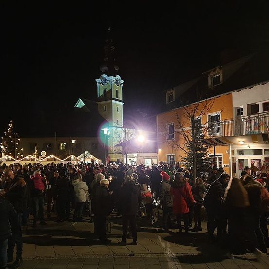 Bei Nacht versammelt sich eine Menschenmenge auf dem Marktplatz für eine Veranstaltung. Der Platz ist von hellen Lichtern und einem hohen Turm mit einer Uhr erleuchtet. Die Menschenmenge ist von Gebäuden mit Weihnachtsdekoration umgeben.