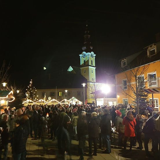 Eine große Menschenmenge versammelt sich bei Nacht draußen, möglicherweise für ein Festival. Es gibt Gebäude mit Weihnachtsdekoration. Ein Turm mit einer Uhr ist in der Ferne sichtbar.