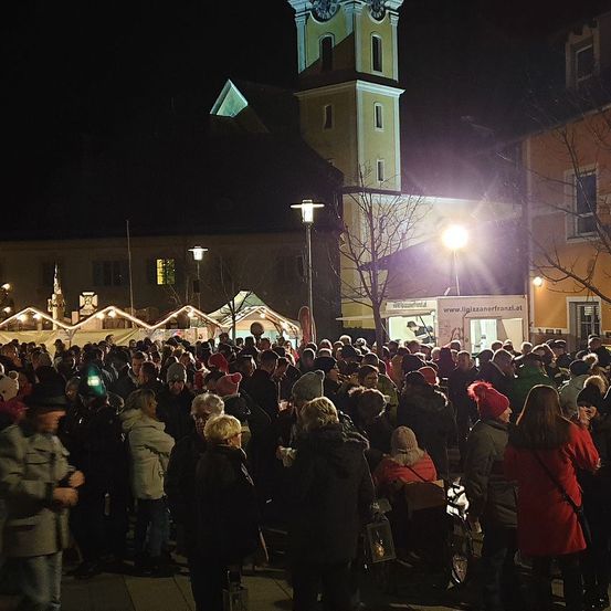 In der Nacht versammelt sich eine Menschenmenge vor einer Kirche mit einem hohen Glockenturm. Einige tragen Mützen und haben Taschen dabei, während andere Taschenlampen benutzen. Ein Imbisswagen und Straßenlaternen sind in der Nähe.