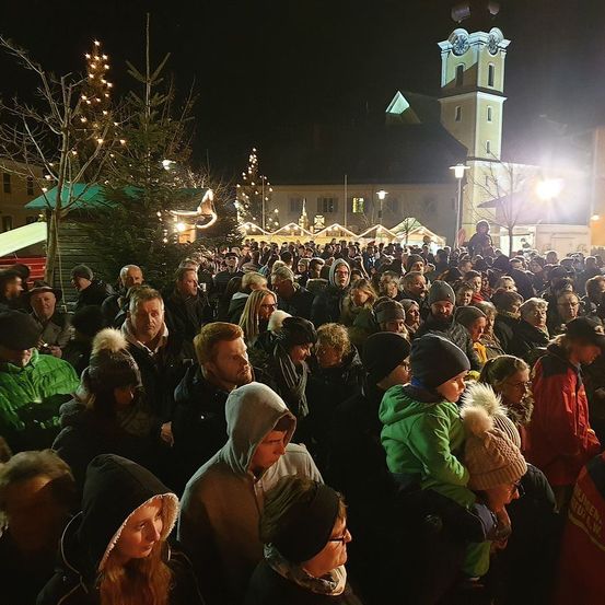 Eine nächtliche Versammlung von Menschen auf einem Stadtplatz, geschmückt mit Weihnachtsbäumen und festlichen Lichtern, mit einem Glockenturm im Hintergrund.