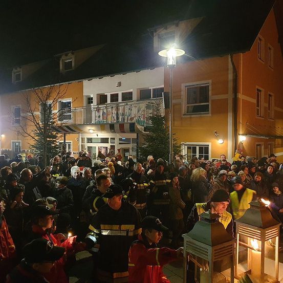 Eine große Menschenmenge versammelt sich bei Nacht vor einem Gebäude, einige halten brennende Laternen. Feuerwehrleute sind anwesend, und das Gebäude wird von Straßenlaternen beleuchtet.