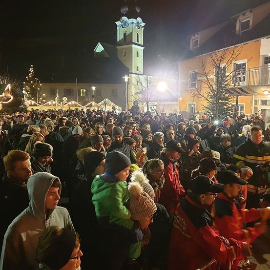 Eine Menschenmenge versammelt sich bei Nacht auf einem Platz in der Stadt, mit einem Uhrturm und einem Weihnachtsbaum im Hintergrund. Viele tragen Winterkleidung und Mützen.