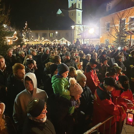 Bei Nacht versammelt sich eine große Menschenmenge im Freien, möglicherweise für eine Feier. Viele tragen Winterkleidung. Einige halten Laternen und andere machen Fotos. Gebäude mit Weihnachtsdekorationen und einem Turm sind sichtbar.