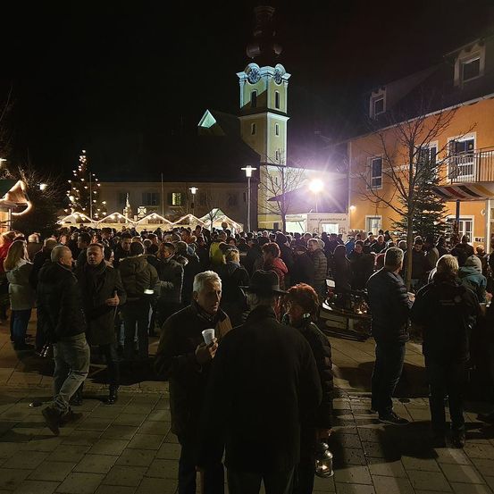 Eine Menschenmenge versammelt sich bei Nacht auf einer Straße, beleuchtet von Straßenlaternen, in der Nähe eines Uhrturms und Gebäuden. Einige tragen Mützen und Mäntel und halten Tassen und Laternen.