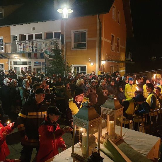 In der Nacht versammelt sich eine Menschenmenge vor einem Gebäude mit vielen Fenstern. Sie halten Kerzen, und zwei große Laternen sind auf einem Tisch beleuchtet. Eine Person steht vor den Laternen.