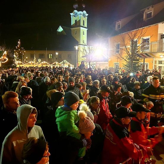 Eine Menschenmenge versammelt sich bei Nacht in einer festlichen Atmosphäre im Freien, mit Gebäuden, Straßenlaternen und einem Turm im Hintergrund. Viele sind warm gekleidet, einige halten Getränke in der Hand.
