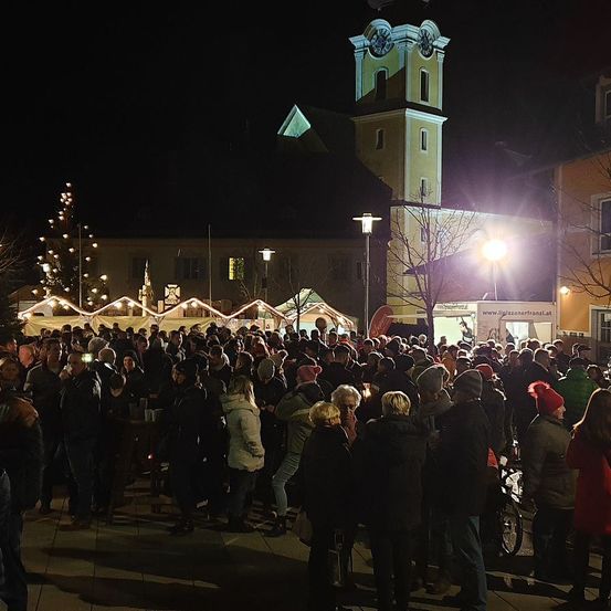 Eine Menschenmenge versammelt sich nachts auf einem Stadtplatz. Hinter ihnen steht ein Glockenturm, und festliche Lichter erhellen den Bereich. Viele Menschen tragen Mützen und Jacken, und einige halten Kerzen. Der Stadtplatz ist von Gebäuden umgeben und mit Weihnachtsbäumen geschmückt.