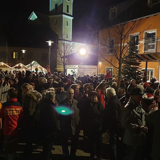 Eine Menschenmenge versammelt sich nachts auf einem Stadtplatz. Sie tragen Winterkleidung und einige haben Mützen auf. Vor einem Gebäude steht ein Baum mit einer Weihnachtsdekoration.