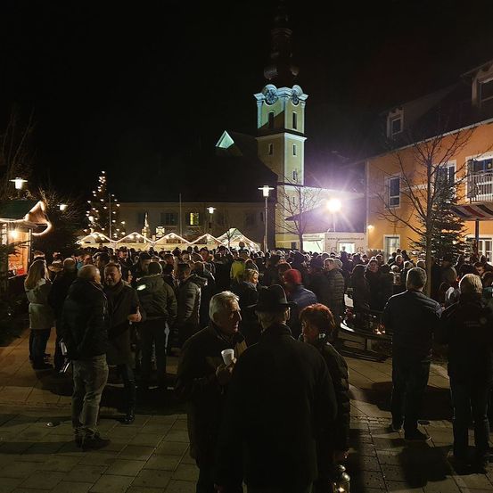 Eine große Menschenmenge steht bei Nacht auf einer Straße. Einige halten Tassen. Im Hintergrund befindet sich ein hoher Glockenturm, und viele Straßenlaternen sind beleuchtet.