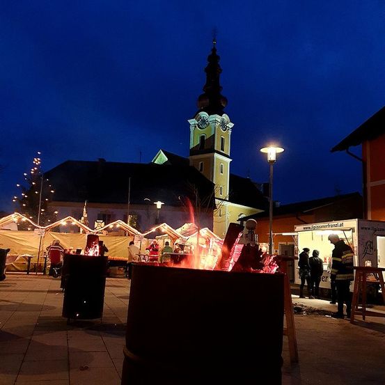 Bei Dämmerung ist auf einem Stadtplatz ein kleiner Markt aufgebaut, mit einem Glockenturm im Hintergrund. Feuerstellen sorgen für Wärme.