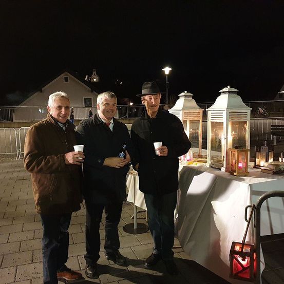 Drei Männer stehen bei Nacht zusammen, halten Tassen und Flaschen, vor ihnen steht ein Tisch mit weißer Tischdecke und Laternen.