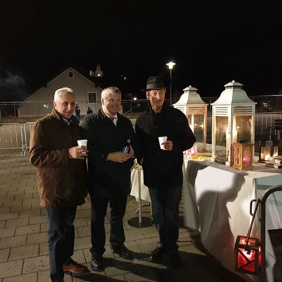 Drei Männer stehen nachts im Freien und lächeln für die Kamera. Sie halten jeweils einen Becher und eine Flasche in der Hand. Hinter ihnen steht ein Tisch mit brennenden Laternen. In der Ferne sind ein Gebäude und ein Fahrrad zu sehen.