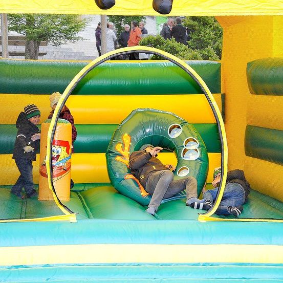 Drei Kinder spielen in einem gelben und grünen aufblasbaren Hüpfburg. Eines kriecht durch ein Loch, während ein anderes auf einem grünen Ring sitzt. Draußen stehen mehrere Leute und beobachten.