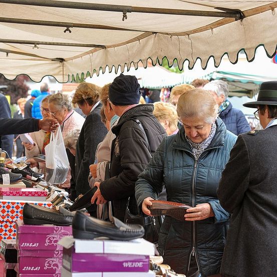 Menschen kaufen Schuhe auf einem Outdoor-Markt unter einem Zelt. Eine Frau betrachtet einen Schuh, während andere in der Nähe stöbern.