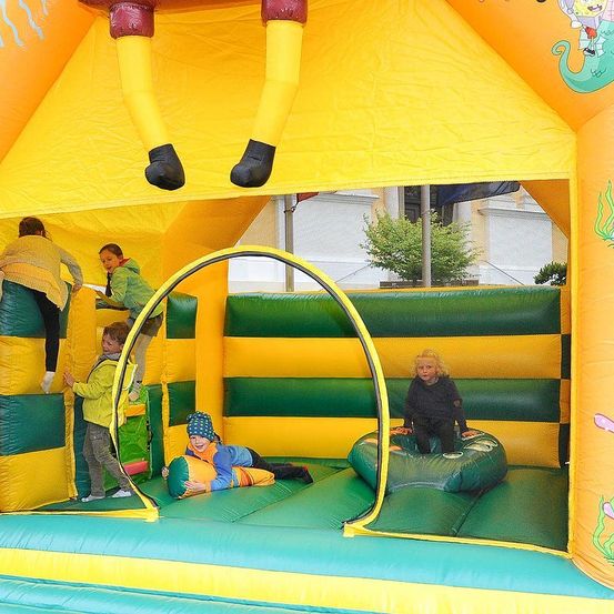 Mehrere Kinder spielen in einem großen aufblasbaren Spielbereich mit gelbem und grünem Thema. Einige klettern, während andere auf dem Aufblasbaren sitzen. Der Aufblasbare hat einen großen, comicartigen Charakter an seiner Seite.