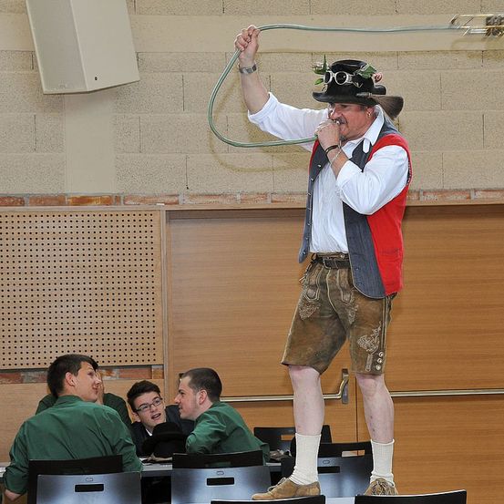 Ein Mann in traditioneller bayerischer Kleidung, einschließlich eines Huts mit Brille und Federn, steht auf einem Stuhl und scheint eine Aufführung zu geben oder zu einem Publikum zu sprechen. Im Hintergrund sitzen mehrere Personen an einem Tisch.