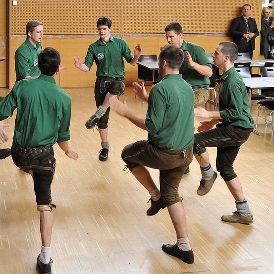 Eine Gruppe junger Männer in grünen Hemden und schwarzen Shorts führt einen traditionellen Tanz in einem Raum auf. Sie sind barfuß oder tragen Socken.