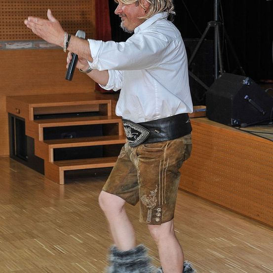 Ein Mann in traditioneller deutscher Kleidung tritt auf der Bühne auf, hält ein Mikrofon und hat einen Lautsprecher und eine Treppe im Hintergrund.
