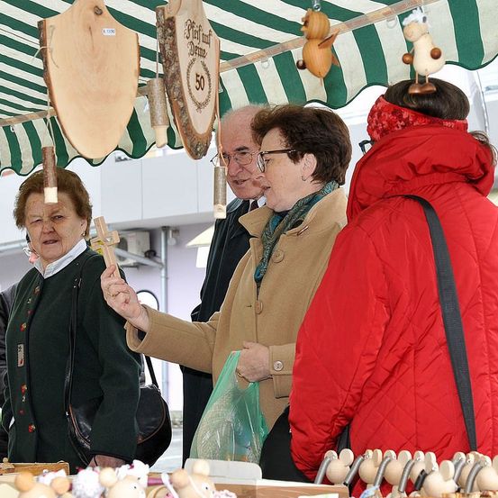 Eine Frau mit Brille hält ein Holzspielzeug und zeigt es einer anderen Frau. Ein Mann in einem schwarzen Anzug steht hinter ihnen. Sie stehen unter einem grün-weißen Vordach. Holzspielzeuge hängen am Vordach.