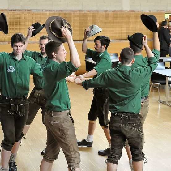 Eine Gruppe von Männern, die grüne Hemden und braune Hosen tragen, tanzt gemeinsam in einem Saal und hält Hüte.