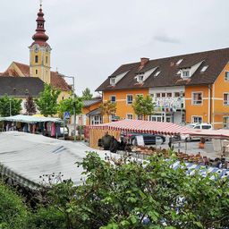 Ein Stadtplatz mit einem Marktstand, der Hüte und Kleidung verkauft, einem gelben Gebäude mit vielen Fenstern und einem Turm mit einer Uhr im Hintergrund.