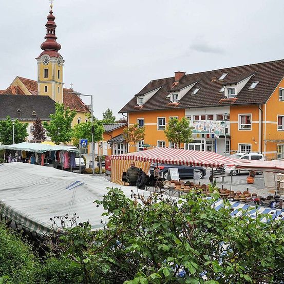 Ein Stadtplatz mit einem Marktstand, der Hüte und Kleidung verkauft, einem gelben Gebäude mit vielen Fenstern und einem Turm mit einer Uhr im Hintergrund.