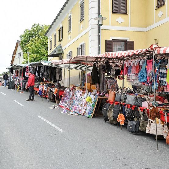 Ein Straßenmarkt mit verschiedenen Ständen, die Kleidung, Handtaschen und andere Artikel verkaufen, ist am Straßenrand vor einem gelben Gebäude aufgebaut.