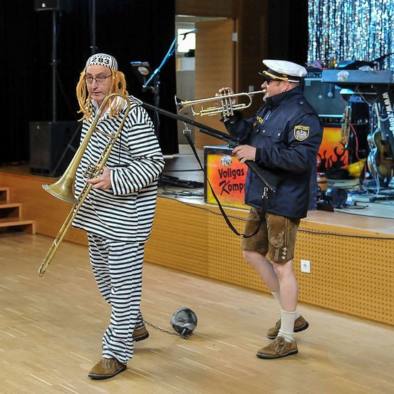 Zwei Männer treten auf der Bühne auf; einer spielt Trompete, der andere Posaune. Sie tragen thematische Kostüme, einer gekleidet als Gefangener. Ein Banner steht 'Vollgas Komp'.