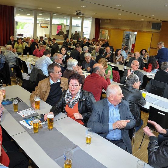 Ein großer Raum mit vielen Menschen, die an Tischen mit Biergläsern sitzen. Die Menschen klatschen und scheinen eine gute Zeit zu haben.