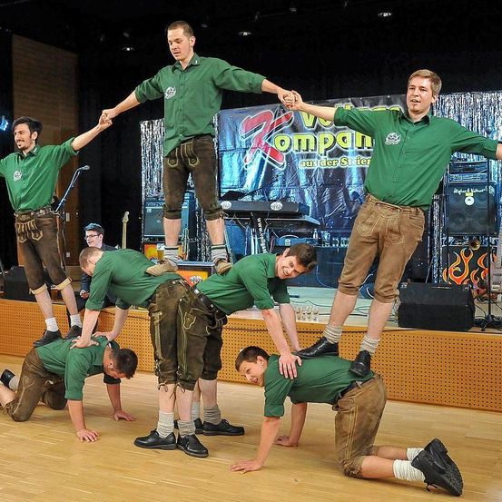 Eine Gruppe von Männern führt einen Balanceakt auf der Bühne auf. Sie sind in grünen Hemden und braunen Hosen gekleidet. Sie stehen auf den Schultern und Händen der anderen. Ein Banner mit dem Text Kompani aus der Stirn hängt im Hintergrund.