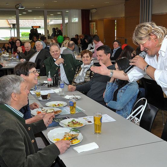 Ein Mann mit Mikrofon spricht zu einem sitzenden Publikum in einem Speisesaal, die Leute lachen und essen, einige tragen Brillen und Krawatten.