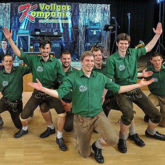 Eine Gruppe von Männern in grünen Hemden und braunen Hosen tritt auf einer Bühne auf, wobei ein Mann kniet und andere hinter ihm stehen.