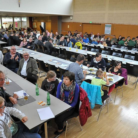 Eine große Gruppe von Menschen sitzt in einem Speisesaal an Tischen, einige essen, während andere in Gespräche vertieft sind. Die Tische sind ordentlich angeordnet und mit weißen Tischdecken bedeckt. Flaschen und Gläser stehen auf den Tischen.