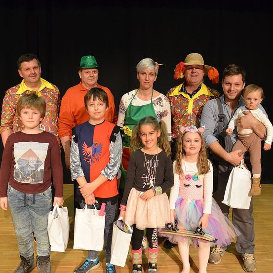 Eine Gruppe von Menschen steht zusammen auf einer Bühne, gekleidet in farbenfrohe Kostüme. Einige tragen Hüte und andere halten Taschen. Die Gruppe besteht aus Erwachsenen und Kindern, die alle lächeln und nach vorne schauen. Hinter ihnen befindet sich ein Hintergrund aus schwarzen Vorhängen.