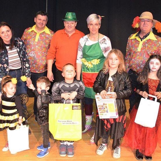 Eine Gruppe von Menschen in verschiedenen Kostümen posiert für ein Foto, darunter eine Frau in einem Blumenshirt, ein Mann in einem Batman-Kostüm und ein Mädchen in einem Hexenkostüm. Sie alle halten Taschen.