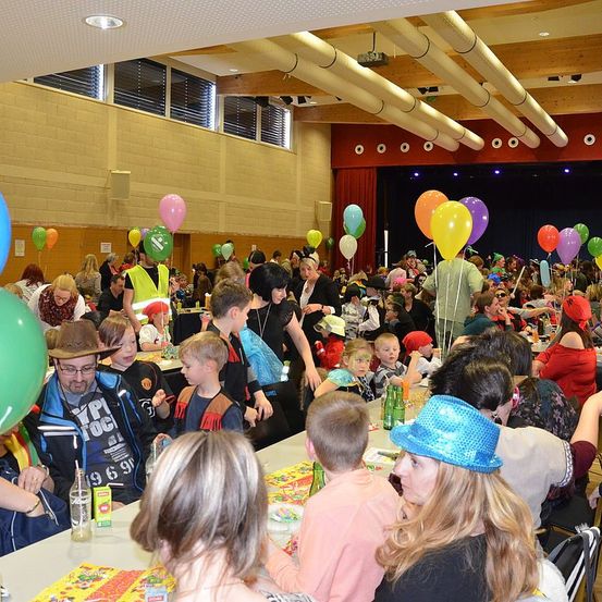 Eine große Gruppe von Menschen, darunter Kinder und Erwachsene, sitzen um Tische in einem Saal. Einige tragen Hüte und Brillen. Viele bunte Luftballons schweben in der Luft. Der Saal hat eine Bühne mit rotem Vorhang und einem Projektor.