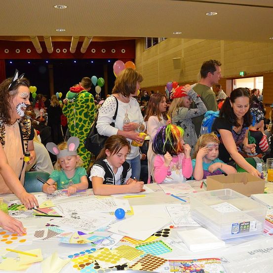 In einem großen Raum sind Kinder um einen Tisch mit Malbüchern und Materialien versammelt. Einige tragen Gesichtsbemalung und Hüte. Erwachsene sind anwesend, einschließlich einer Frau mit einem Getränk. Der Raum hat Luftballons und eine Bühne mit einem Vorhang.