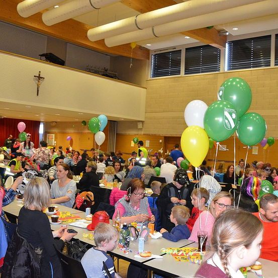 In einem großen Raum mit Tischen und Stühlen sitzen eine Vielzahl von Menschen, darunter Erwachsene und Kinder. Sie essen und genießen eine festliche Atmosphäre, mit bunten Luftballons und Dekorationen, die von der Decke hängen.