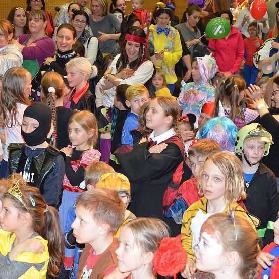 Eine Menschenmenge aus Kindern und Erwachsenen in Kostümen bei einer Veranstaltung, einige halten Luftballons und tragen Schminkgesichter. Die Kinder sehen aufgeregt und engagiert aus, warten möglicherweise auf eine Ankündigung.