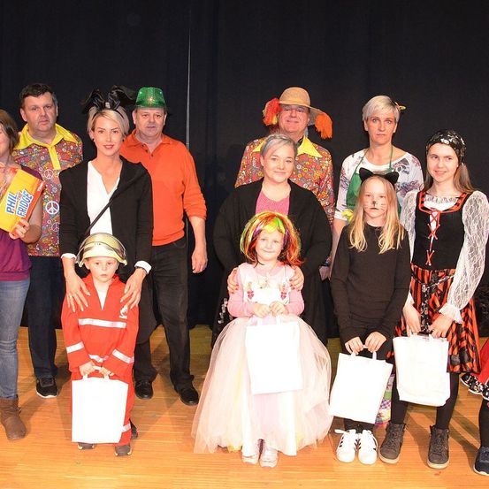Eine Gruppe von Erwachsenen und Kindern in Kostümen posiert für ein Foto auf einer Bühne. Einige halten weiße Taschen. Die Kinder tragen Gesichtsbemalung.