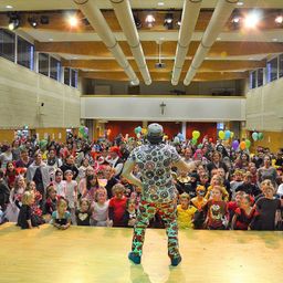 Ein Saal voller Menschen, einschließlich Kinder in Kostümen, die einen Mann in einem gemusterten Outfit beobachten. Der Mann steht auf der Bühne, mit farbenfrohen Luftballons in der Luft. Der Saal hat Holzbalken an der Decke und weiße Wände.