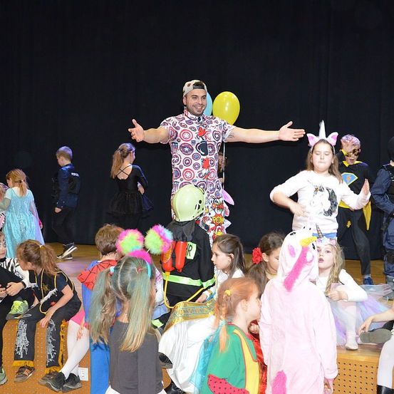 Ein Mann in einem farbenfrohen Kostüm steht auf der Bühne, umgeben von Kindern. Einige Kinder tragen Kostüme, darunter ein Feuerwehrmann und ein Einhorn. Der Mann hält einen Ballon.