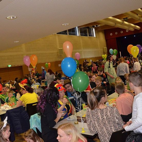 Eine große Menschenmenge, einschließlich Kinder, Erwachsene und Personen mit Partyhüten, versammelt sich in einem Raum mit Luftballons, Tischen und Stühlen. Einige Leute essen, während andere stehen und reden. Der Raum hat eine hohe Decke mit Lichtern und einem offenen Fenster.