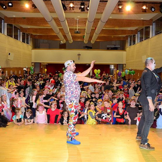 Ein Saal voller Kinder in Kostümen, die zwei Männer auf der Bühne beobachten, einer zeigt mit dem Finger und der andere sieht zu, mit Luftballons, die herumschweben.