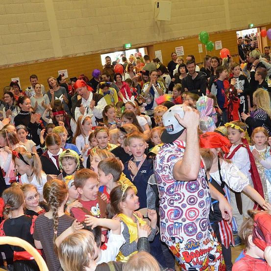 Eine große Menschenmenge aus Kindern und Erwachsenen in Kostümen versammelt sich in einer Halle. Viele jubeln und klatschen, während einige Masken tragen. Bunte Luftballons und Schilder schmücken die Wände.