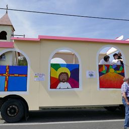 Ein dekorierter Lastwagen mit Jesus in der mittleren Fensterscheibe gemalt, neben einer Kirche geparkt. Menschen stehen auf dem Dach des Lastwagens, und ein Mann steht neben ihm.
