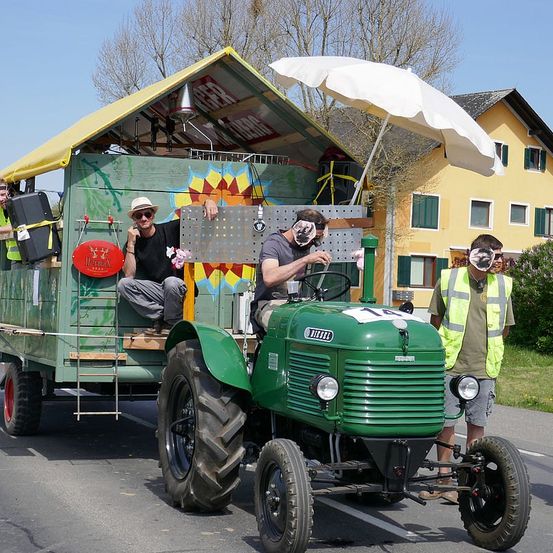 Zwei Männer sitzen auf einem grünen Traktor mit gelbem Dach. Sie tragen Masken und Sonnenbrillen. Ein Regenschirm ist am Traktor befestigt. Eine Person steht neben dem Traktor. Ein Gebäude mit Balkon befindet sich hinter dem Traktor.