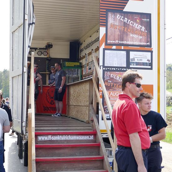 Ein Lastwagen ist am Straßenrand geparkt. Zwei Männer stehen auf den Treppen des Trucks. Ein anderer Mann schaut sie an.